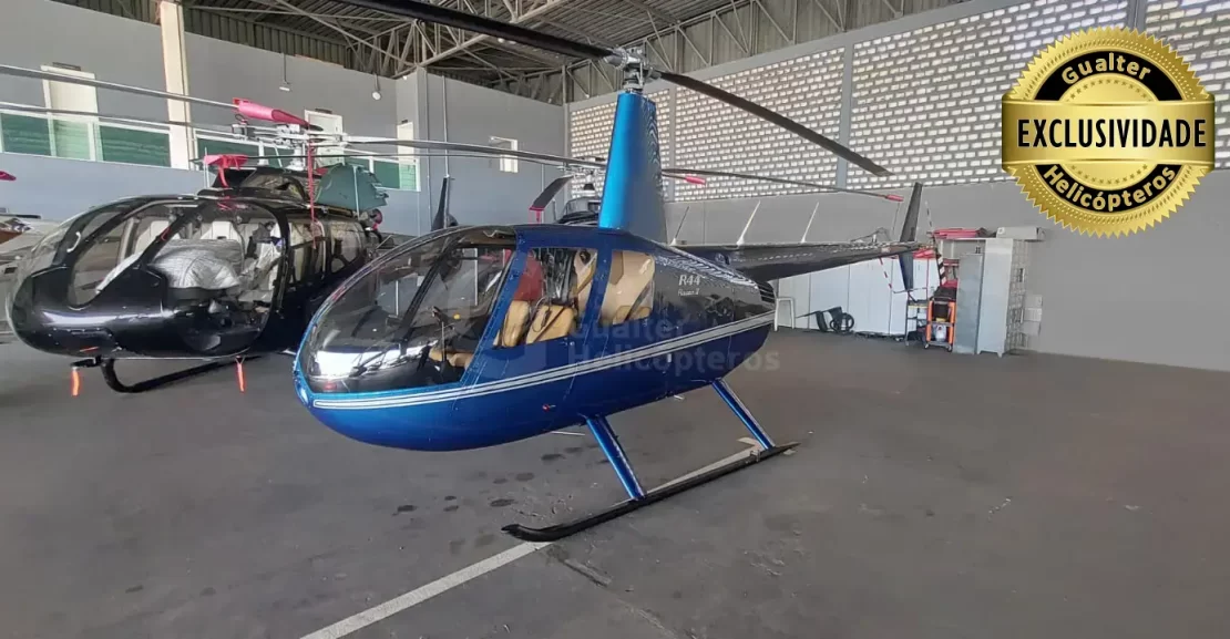 Helicóptero Robinson R44 Raven II ano 2010 com 7.343 horas totais de voo à venda na Gualter Helicópteros. Aeronave azul com listras branca lateral com bancos de couro em bege. O helicóptero está pousado em um hangar coberto e a foto foi tirada na diagonal de frente.