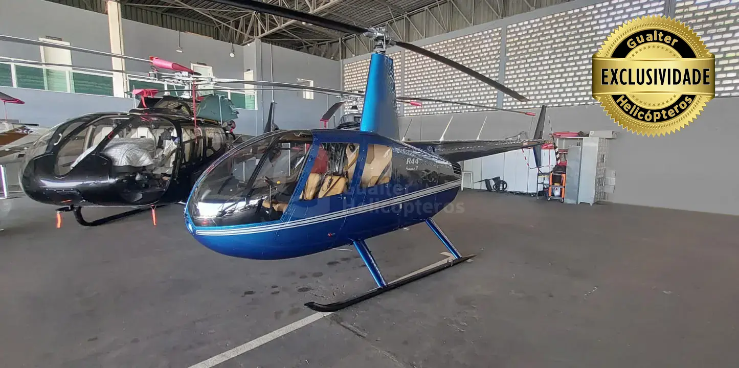 Helicóptero Robinson R44 Raven II ano 2010 com 7.343 horas totais de voo à venda na Gualter Helicópteros. Aeronave azul com listras branca lateral com bancos de couro em bege. O helicóptero está pousado em um hangar coberto e a foto foi tirada na diagonal de frente.