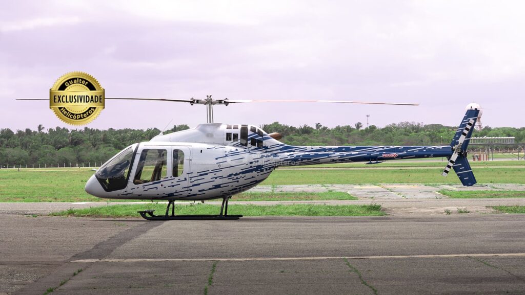 Bell 505 prata com detalhes em azul pousado. Selo de exclusividade na parte superior esquerda da imagem e logo da Gualter Helicópteros ao centro como marca d'água. Aeronave usada à venda na Gualter Helicópteros.