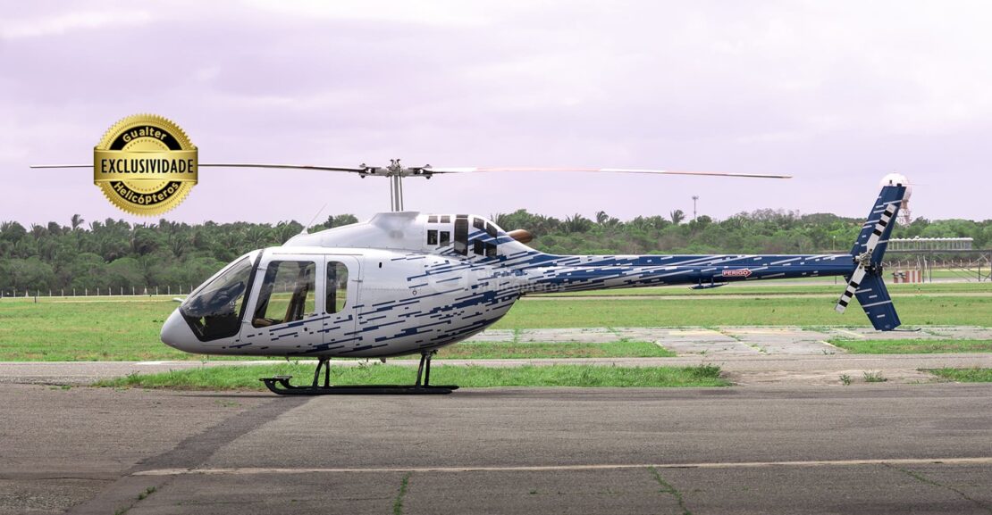 Bell 505 prata com detalhes em azul pousado. Selo de exclusividade na parte superior esquerda da imagem e logo da Gualter Helicópteros ao centro como marca d'água. Aeronave usada à venda na Gualter Helicópteros.