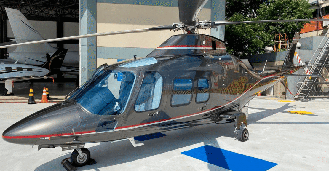 Helicóptero AgustaWestland A109S Grand 2010 grafite com faixa vermelha, visto de frente e lado esquerdo, pousado.